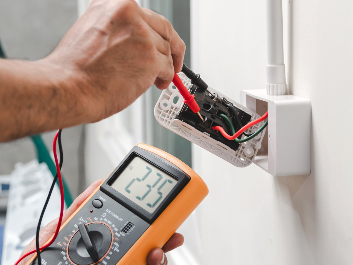 A person uses a multimeter to measure electrical voltage in a wall socket, showing a reading of 235 volts.