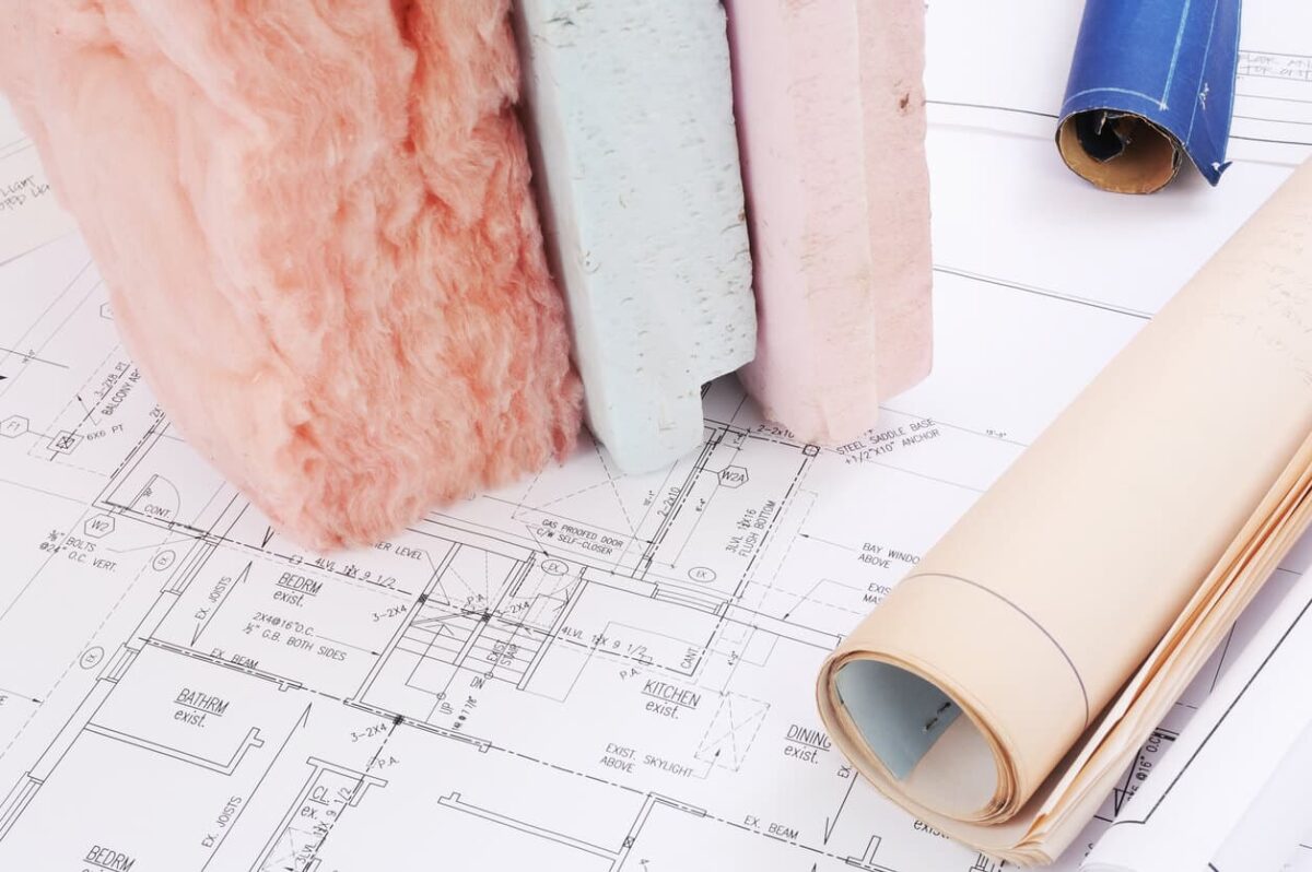 Rolls of blueprints and samples of insulation materials, including pink fiberglass and foam boards, are displayed on a drafting table.