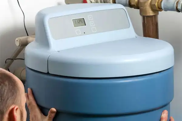 A person meticulously inspects a large blue water softener with a digital display, surrounded by intricate plumbing pipes, ensuring flawless water softener installation.