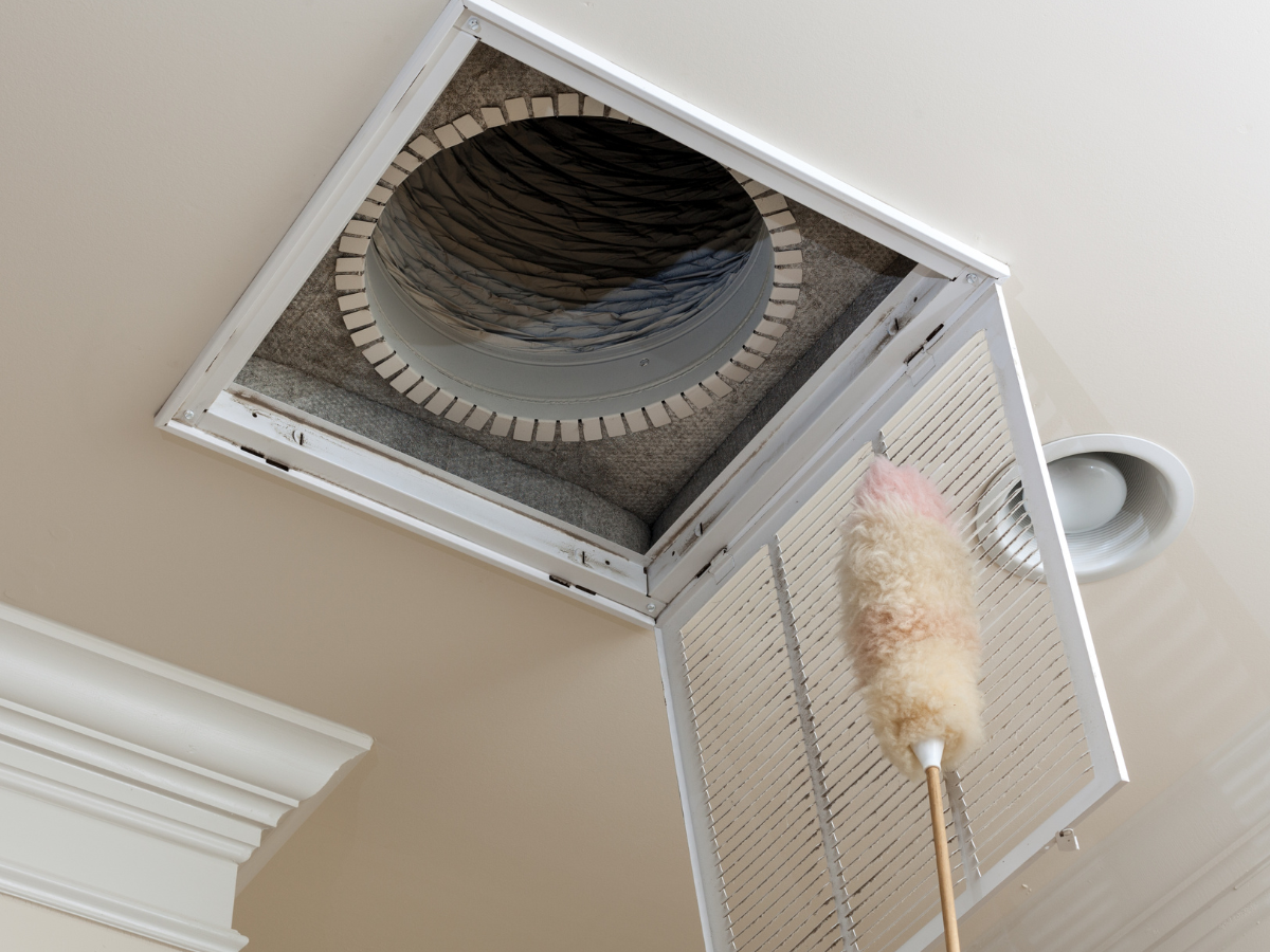 An open ceiling air vent is being cleaned with a feather duster, revealing the ductwork inside.