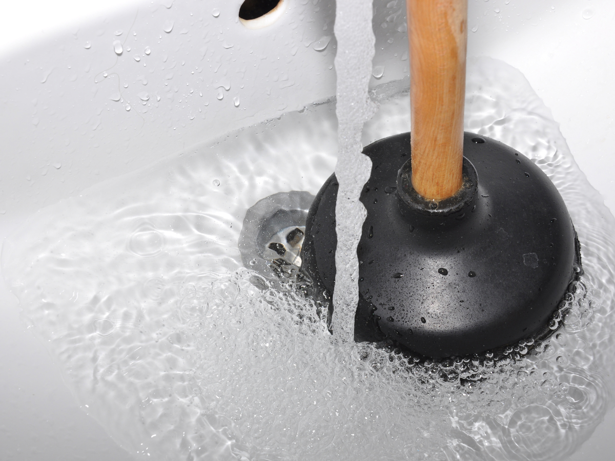 A plunger with a wooden handle is placed in a white sink, surrounded by running water.