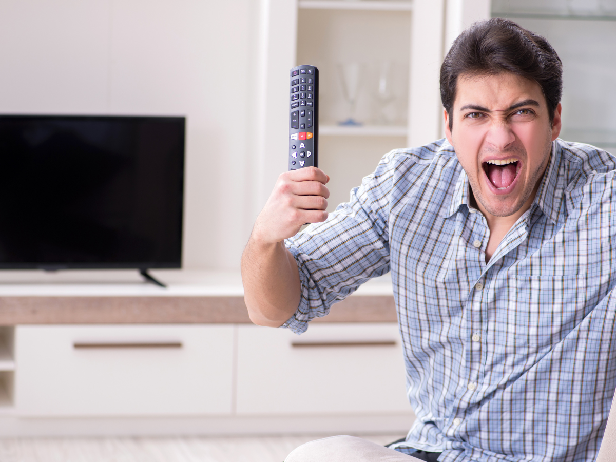 A man in a plaid shirt excitedly holds a TV remote in front of a television.