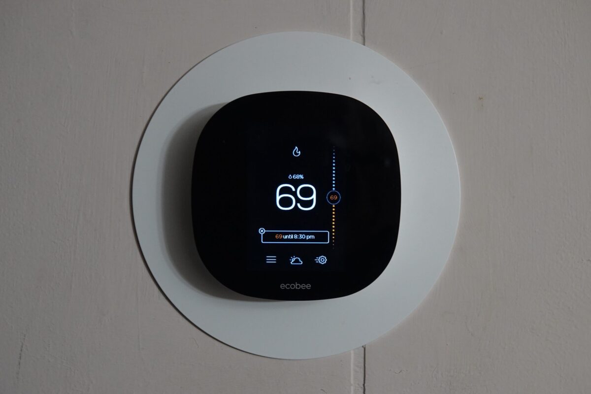 A digital thermostat mounted on a wall displays the number 69. The device screen shows buttons and settings on a circular interface.