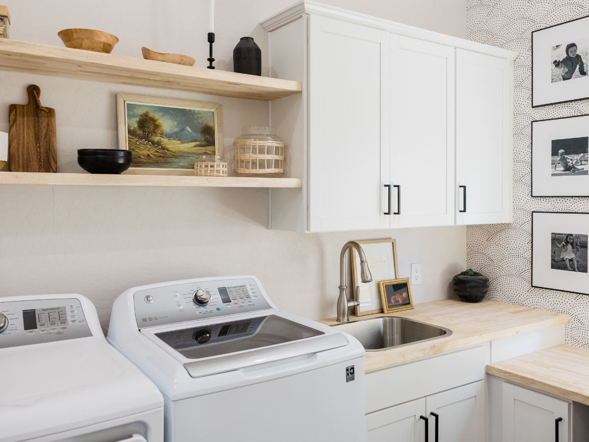Bright laundry room with white cabinets, wooden countertops, washer, dryer, and a sink. Open shelves hold decor items like a painting, basket, and cutting boards. Black-and-white photos on wall.