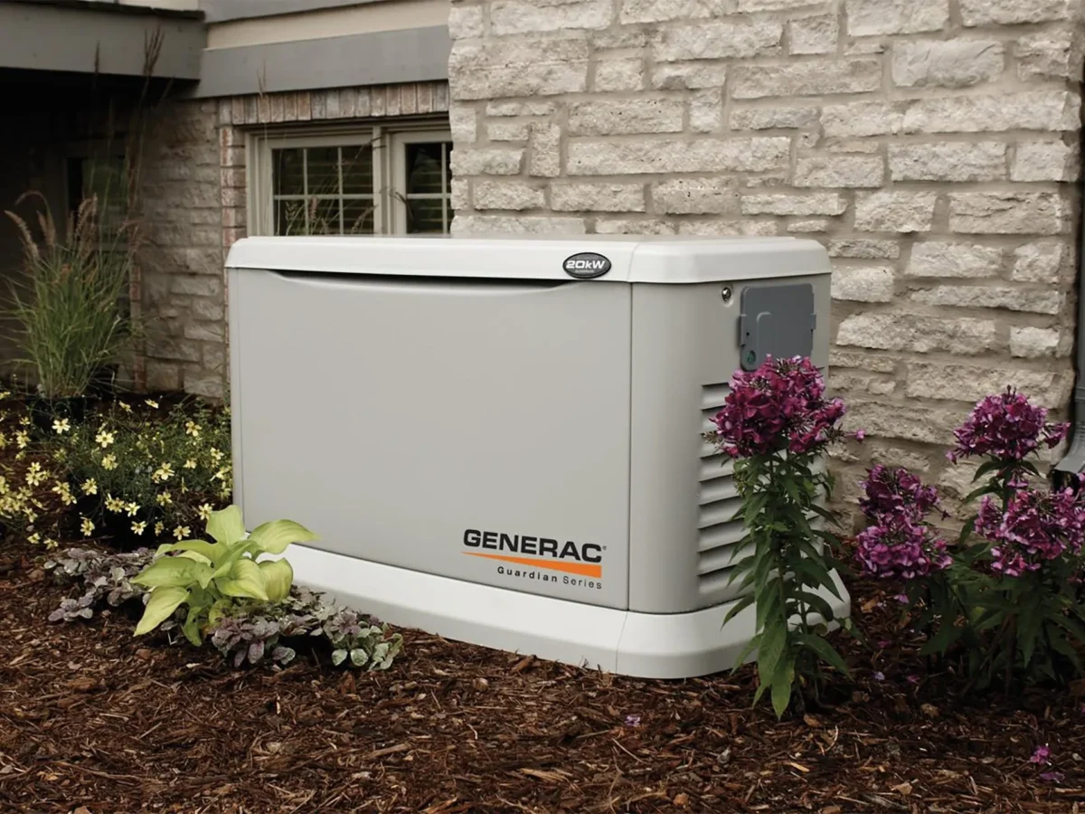 A Generac Guardian Series standby generator is installed next to a brick house, surrounded by flowers and mulch, offering peace of mind like reliable water softener repair services ensuring smooth household operations.