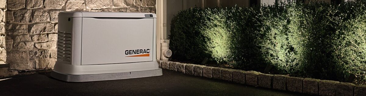 A Generac generator is installed on a paved area beside a stone wall and illuminated shrubs in a nighttime setting.