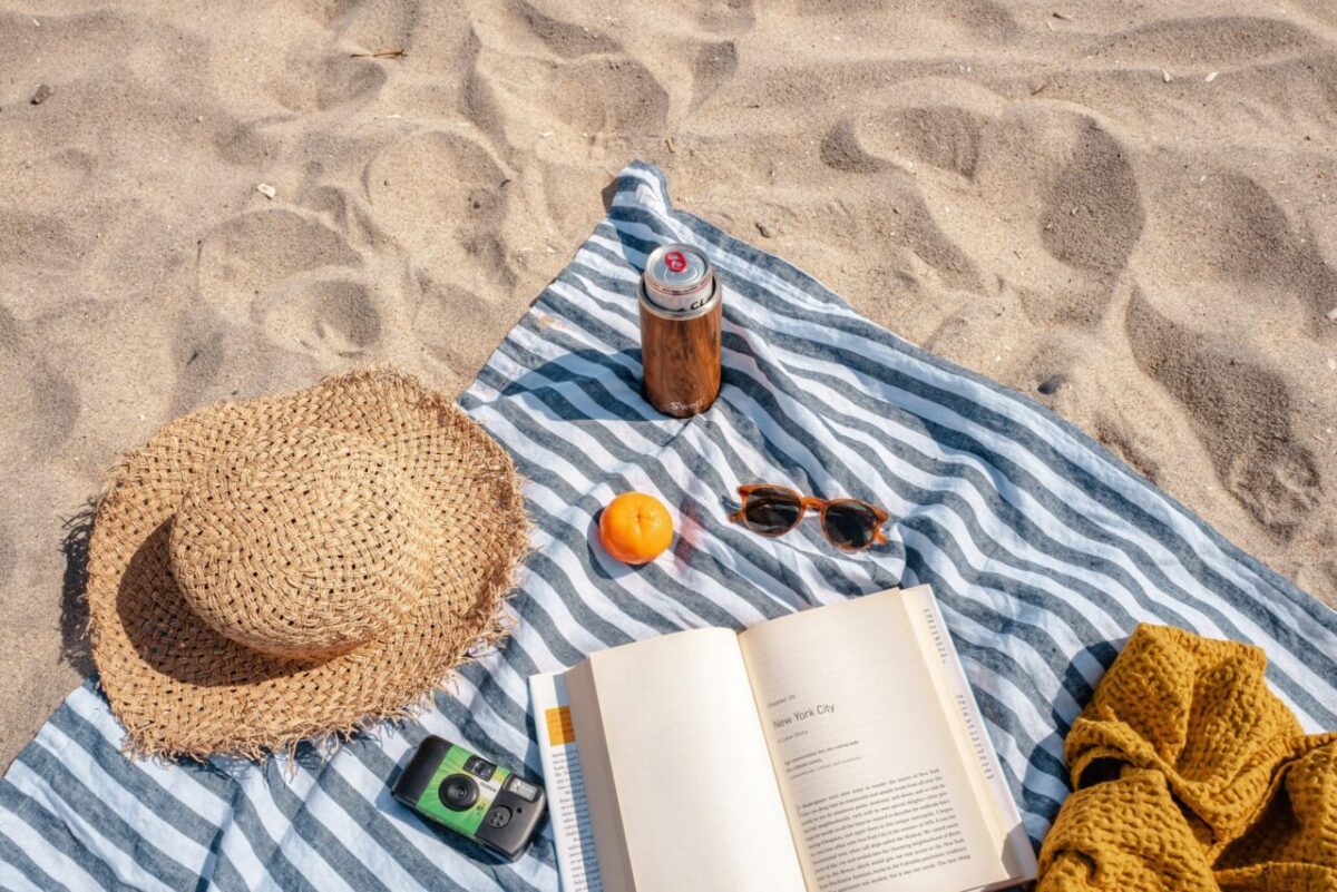 A straw hat, canned drink, orange, sunglasses, camera, and open book are arranged on a striped blanket on sandy beach.