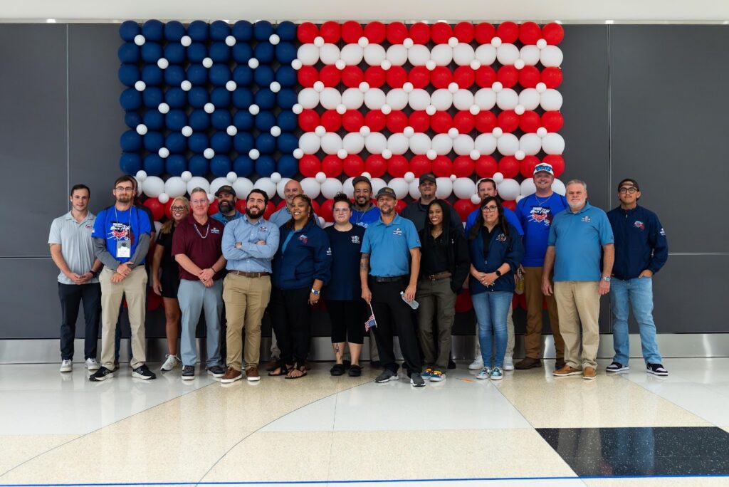 A group of people pose in front of a wall decorated with red, white, and blue balloons arranged to resemble the American flag.