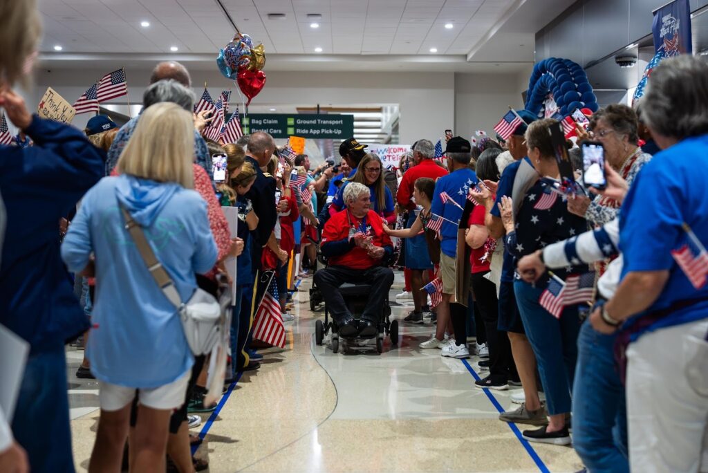 A crowd lines an airport corridor, many holding American flags, as an elderly person in a wheelchair moves through, greeted and photographed by onlookers.