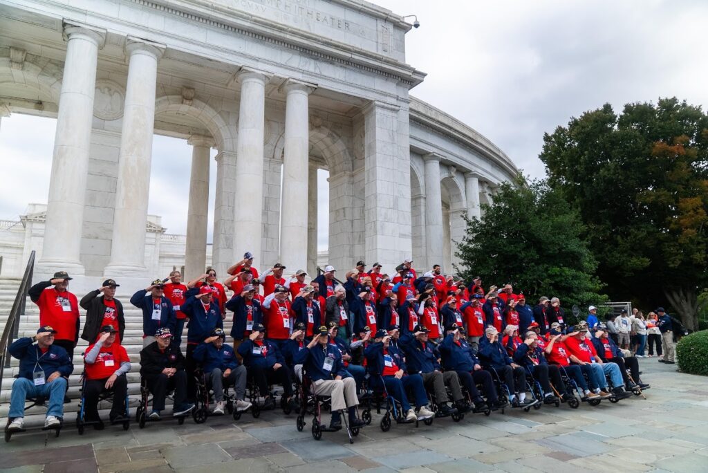 A large group of veterans, many in wheelchairs, pose for a photo in front of a white marble monument with columns.