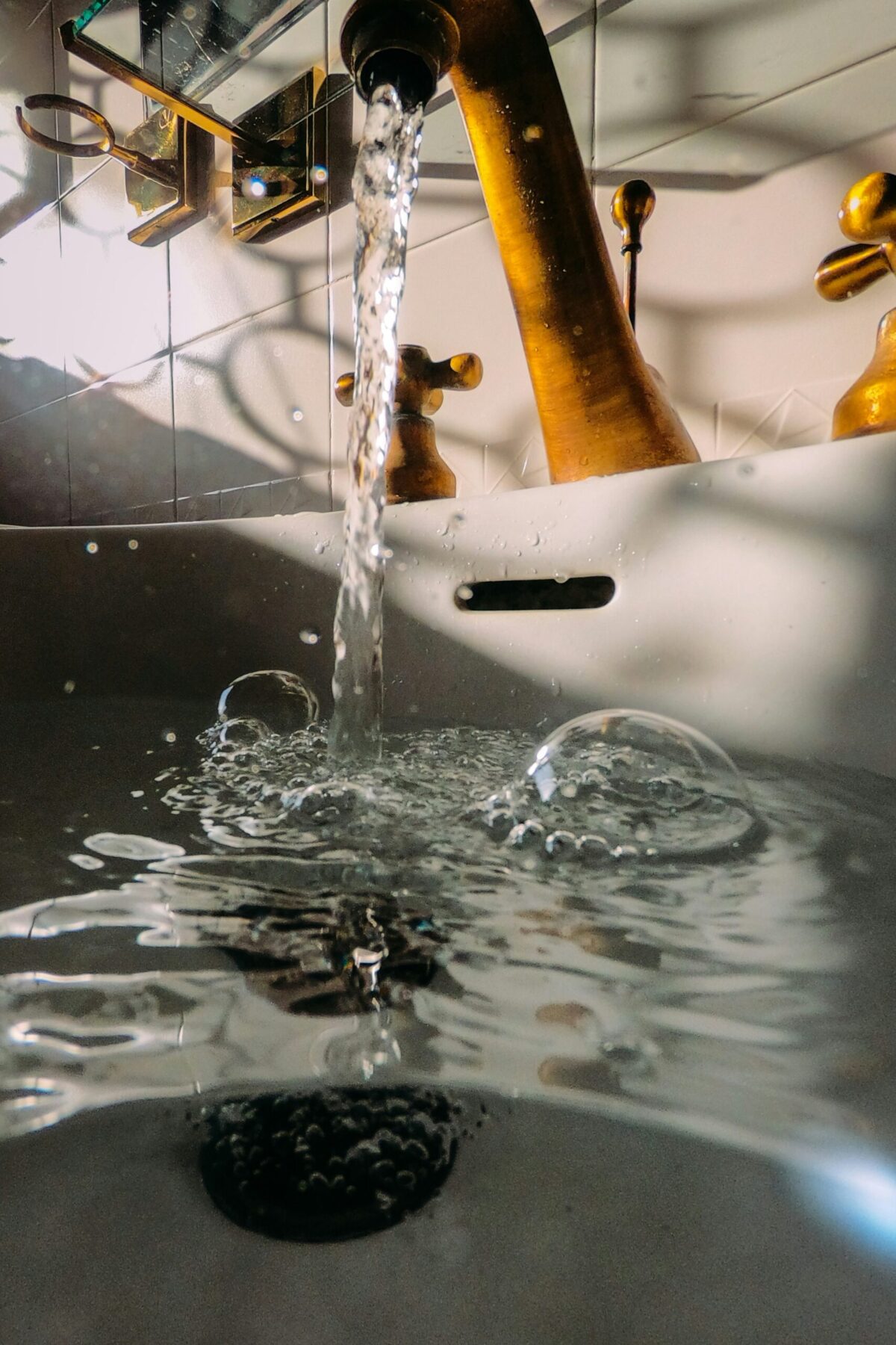 Water flows from a brass faucet into a white sink, creating bubbles and ripples on the water's surface below.