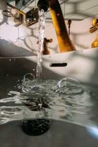 Water flows from a brass faucet into a white sink, creating bubbles and ripples on the water's surface below.