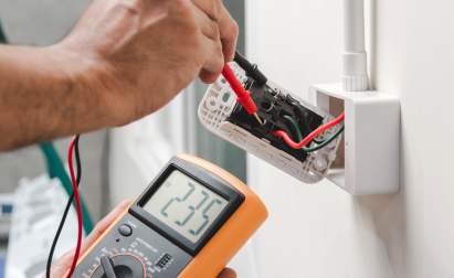 A person uses a multimeter to measure electrical voltage in a wall socket, showing a reading of 235 volts.
