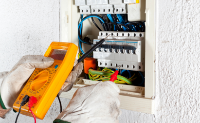 Person using a digital multimeter to measure voltage in an electrical panel. Wearing protective gloves and adjusting wires.