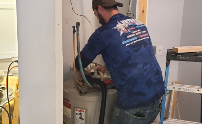 A man in a cap and blue shirt works on a water heater. Tools and a ladder are nearby in a room under maintenance.