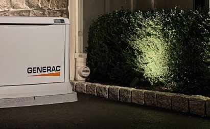 A Generac generator is installed on a paved area beside a stone wall and illuminated shrubs in a nighttime setting.