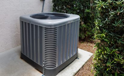 Outdoor air conditioning unit situated on a concrete pad, surrounded by shrubs, with a wall and electrical connection visible in the background.