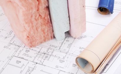Rolls of blueprints and samples of insulation materials, including pink fiberglass and foam boards, are displayed on a drafting table.