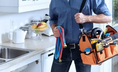 Person holding a bag of plumbing tools and hoses standing in a kitchen with a fruit dish on the counter.