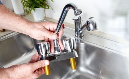 Person holding a detached kitchen faucet over a stainless steel sink, with a potted plant in the background.