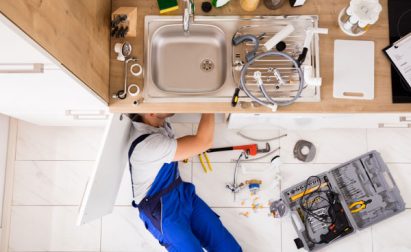 A person in blue overalls lies on the floor under a kitchen sink, surrounded by tools and plumbing parts, working on repairs.
