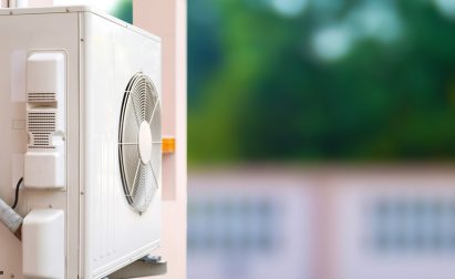 Outdoor air conditioning unit mounted on a wall, with greenery visible in the blurred background.