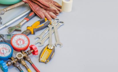 Assorted tools and workshop equipment, including gauges, pliers, wrenches, gloves, and an angle grinder, displayed on a gray surface.