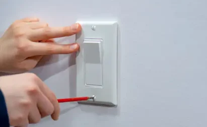 Person using a screwdriver to adjust or install a white light switch on a wall.
