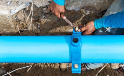 Person in blue shirt tightening bolts on a bright blue pipe using a wrench, with soil and debris around.