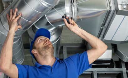 Worker in blue uniform and cap inspects a large metal duct in an industrial setting. He is using a flashlight to examine the structure.