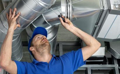 Worker in blue uniform and cap inspects a large metal duct in an industrial setting. He is using a flashlight to examine the structure.