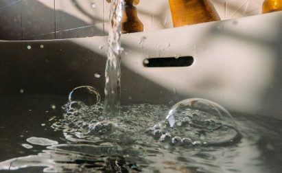 Water flows from a brass faucet into a white sink, creating bubbles and ripples on the water's surface below.
