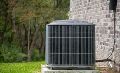 An outdoor air conditioning unit sits on a concrete base next to a brick wall, surrounded by grass and trees in the background.