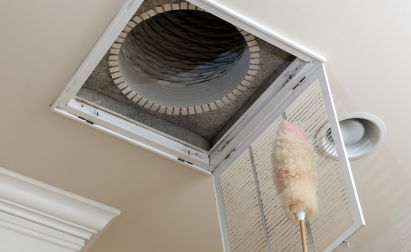 An open ceiling air vent is being cleaned with a feather duster, revealing the ductwork inside.