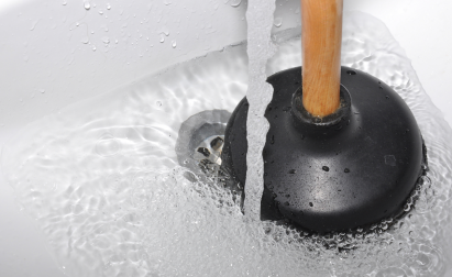 A plunger with a wooden handle is placed in a white sink, surrounded by running water.