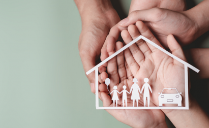 Hands hold paper cutouts of a family and car under a house outline on a soft green background, symbolizing protection and security.