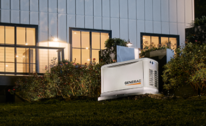 A Generac generator installed outside a house with large windows, surrounded by bushes, under evening light.