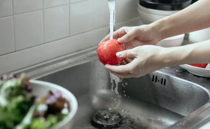 Person washes a red apple under running water in a kitchen sink, with a bowl of vegetables nearby.