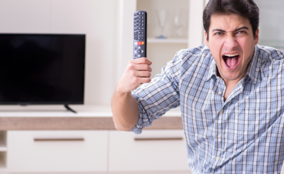 A man in a plaid shirt excitedly holds a TV remote in front of a television.