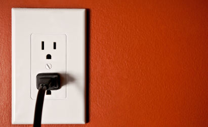 A black electrical plug is inserted into a white wall outlet on a red wall.