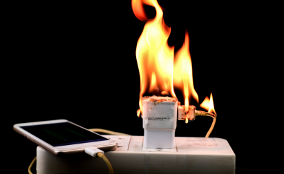 A smartphone charger plugged into an outlet is on fire against a black background.