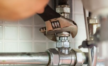 A hand using a wrench to tighten a pipe fitting in a plumbing setup against a tiled wall background.