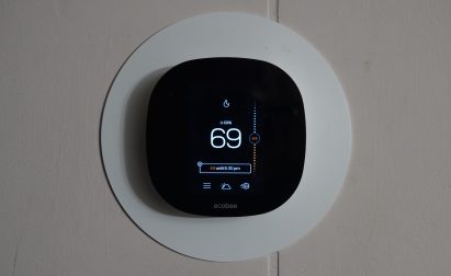A digital thermostat mounted on a wall displays the number 69. The device screen shows buttons and settings on a circular interface.