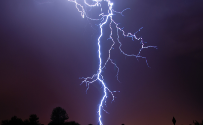 A bright lightning bolt strikes the ground against a dark sky, illuminating surrounding clouds and silhouetting trees below.