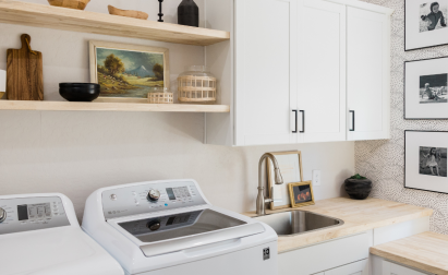 Bright laundry room with white cabinets, wooden countertops, washer, dryer, and a sink. Open shelves hold decor items like a painting, basket, and cutting boards. Black-and-white photos on wall.